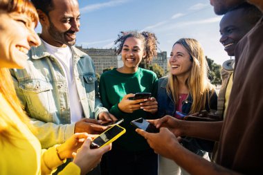 Dışarıda cep telefonu kullanan mutlu, çok ırklı bir grup genç arkadaş. Bağımlı genel z insanlar cep telefonuyla sohbet ve uygulama üzerinde sosyal medya içeriği izlemek
