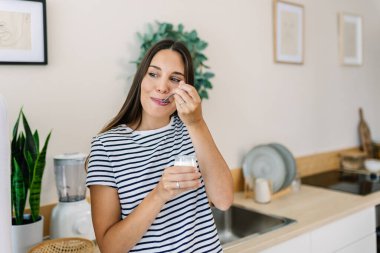 Mutfakta elinde yoğurt kabıyla duran genç bir kadın. Gülümseyen kadın evde sağlıklı bir süt ürününün tadını çıkarıyor. Sağlıklı yaşam tarzı ve refah kavramı.