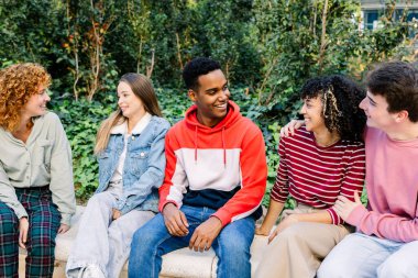 Bir grup genç arkadaş parkta bir bankta oturuyor, günlük kıyafetlerle gülüyor ve sohbet ediyorlar, arkadaşlığı, bağlantıyı ve eğlenceyi yakalayan açık havada takılmanın keyfini çıkarıyorlar.