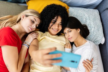 Üç bayan arkadaş bir yatakta uzanıyor, neşeli bir selfie çekerken pijama partisinin tadını çıkarıyorlar, mutluluk ve bağlantının anlarını yakalıyorlar. Kadın arkadaşlığı ve gençlik kavramı