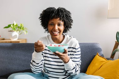 Modern dairesinin oturma odasındaki kanepede oturan sağlıklı kahvaltı kasesi dolusu yoğurt ve meyve yiyen mutlu siyah kadın. Sağlıklı yaşam tarzı ve yeme konsepti
