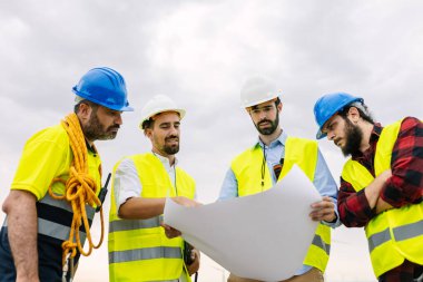 Çeşitli mühendisler ve inşaat işçileri proje planlarını gözden geçirip sürdürülebilir bir mimari plan üzerinde işbirliği yapıyorlar. Yenilenebilir enerji ve endüstriyel işgücü kavramı