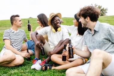 Farklı arkadaş grupları açık havada toplanmaktan zevk alıyor. Şehir parkında çim üzerinde dinlenirken çeşitli genç insanlar biranın tadını çıkarıyor.