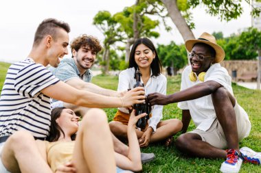 Çok ırklı bir grup genç arkadaş yazın şehir parkında çimlerde dinlenerek eğleniyor.
