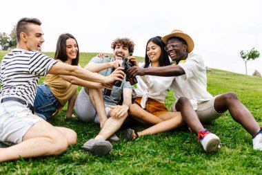 Farklı arkadaş grupları açık havada toplanmaktan zevk alıyor. Şehir parkında çim üzerinde dinlenirken çeşitli genç insanlar biranın tadını çıkarıyor.