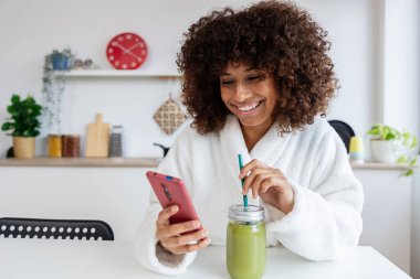 Genç melez, bornozlu mutlu kadın mutfakta yeşil meyve suyu içerken cep telefonu kullanıyor.
