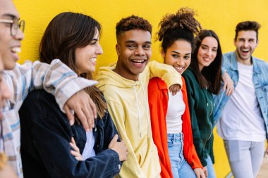 Neşeli farklı arkadaşlar grubu birlikte vakit geçiriyorlar. Parlak sarı bir duvara sarılıp gülüyorlar.