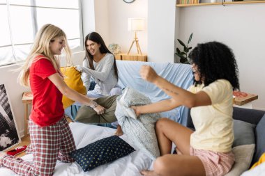 Üç neşeli çok ırklı bayan arkadaş pijama partileri sırasında yastık savaşı yapıyorlar. Kadın arkadaşlığı ve gençlik kavramı