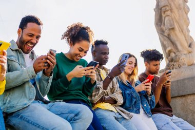 Dışarıda cep telefonu kullanan mutlu, çok ırklı bir grup genç arkadaş. Bağımlı genel z insanlar cep telefonuyla sohbet ve uygulama üzerinde sosyal medya içeriği izlemek