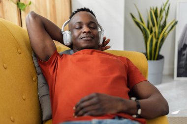 Relaxed African man lying on sofa listening to music with wireless headphones at home. listening to his favorite music and enjoying a peaceful moment at living room. Leisure concept