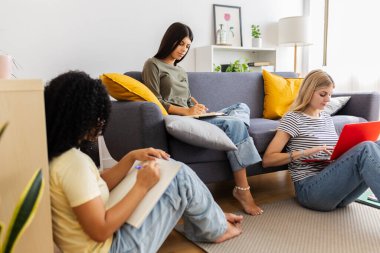 Üç kız öğrenci bir proje üzerinde çalışıyorlar, online araştırmayla rahat bir oturma odası ortamında not almayı birleştiriyorlar. Kadın arkadaşlığı ve gençlik kavramı