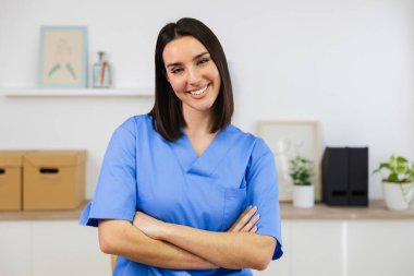 Kollarını kavuşturup gülümseyen genç bir hemşirenin portresi, parlak bir sağlık ofisinde profesyonellik ve sağlık uzmanlığı sergiliyor.