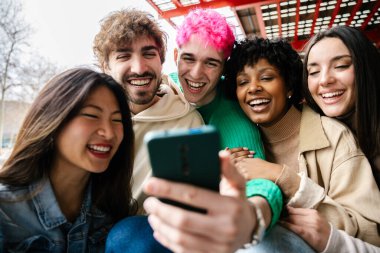 Mutlu arkadaşlar üniversite merdivenlerinde oturup akıllı telefonlardan eğlenceli videolar izliyorlar. Birliktelik, eğlence ve bin yıllık yaşam tarzı konsepti.