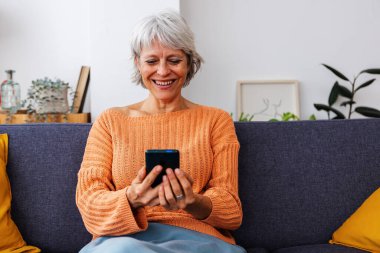 Modern apartmandaki rahat koltukta dinlenirken cep telefonu uygulamasında komik sosyal medya içeriğinin keyfini çıkaran mutlu bir son sınıf kadını. Teknoloji yaşam tarzı konsepti