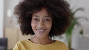 Afro saç stili olan neşeli, Afro-Amerikan bir kadının yakın plan portresi. Kameraya bakıyor ve gülümsüyor. Modern bir oturma odasında gerçek mutluluğu ve olumlu duyguları gösteriyor.