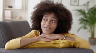 Afro saç stili ile düşünceli genç Afrikalı kadın evdeki kanepede dinleniyor, sakin bir yüz ifadesiyle hayal kuruyor ve gülümsüyor, huzurlu bir yalnızlık anının tadını çıkarıyor.