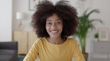 Afro saçlı neşeli Afrikalı bir kadın. Video görüşmesi sırasında mutlu bir şekilde konuşuyor ve el kol hareketi yapıyor. Samimi bir oturma odasındaki kameraya bakıyor.