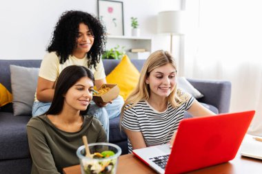 Birlikte yemek yiyen üç kız öğrenci, rahat oturma odalarında dinlenirken mutlu bir şekilde dizüstü bilgisayarlarında geziniyorlar. Kadın arkadaşlığı ve gençlik kavramı