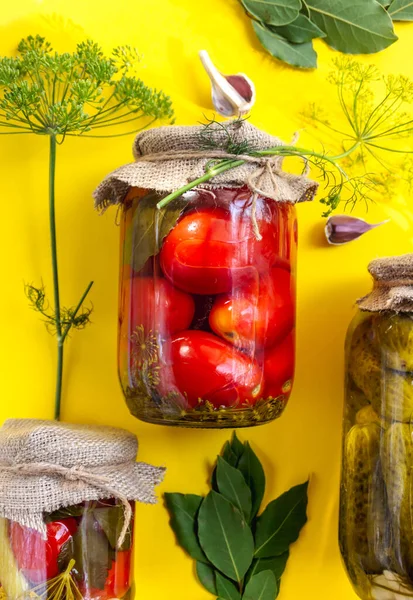 Frascos de vidrio con tomates en escabeche caseros, sellados con tapa metálica sobre fondo amarillo. Pepinos en escabeche, ramitas de eneldo y hojas de laurel —  Fotos de Stock