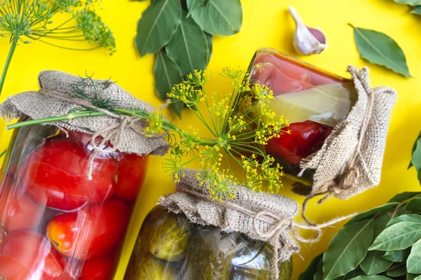 Szklane słoiki z domowej roboty marynowanymi pomidorami, zamknięte metalową pokrywą na żółtym tle. ogórki marynowane, gałązki koperkowe i liście laurowe — Zdjęcie stockowe