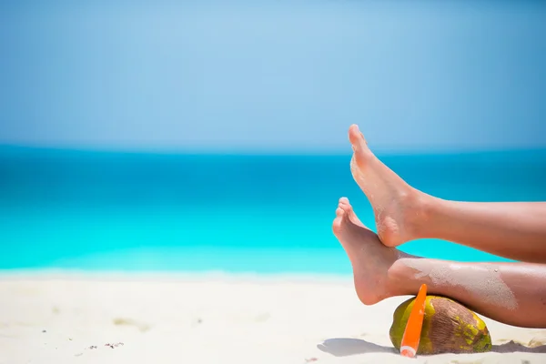 Gambe lisce abbronzate femminili con crema solare e cocco sulla spiaggia bianca — Foto Stock