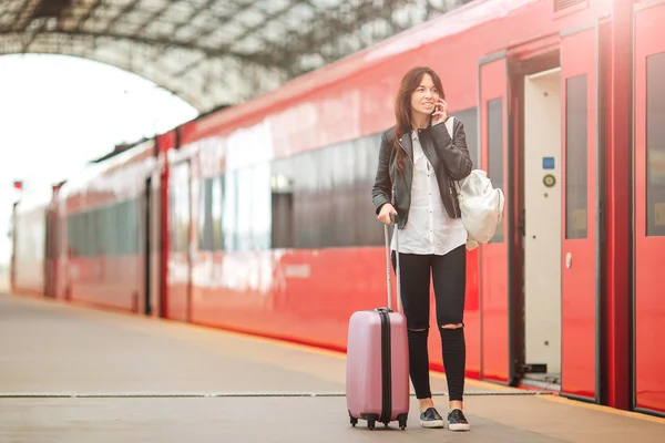 Ung kvinna med bagage pratar via mobiltelefon på en tågstation. Caucasiam turist väntar hennes expresståg medan hon reser. — Stockfoto