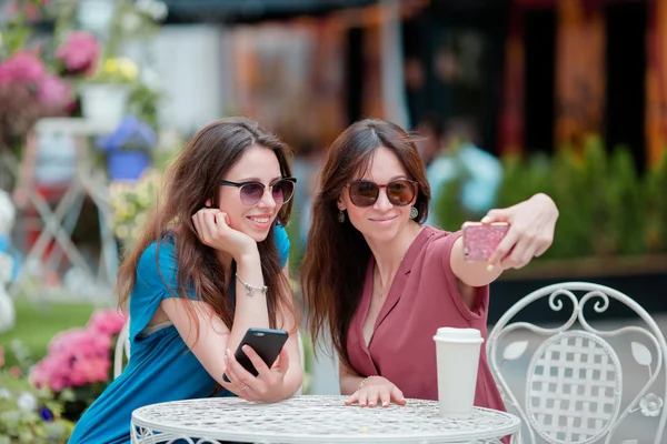 Deux jeunes filles prenant selfie avec téléphone intelligent au café en plein air. Deux femmes après avoir fait leurs courses avec des sacs assis dans un café à ciel ouvert avec du café et en utilisant un smartphone — Photo