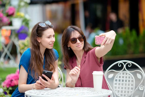 Duas meninas tomando selfie com telefone inteligente no café ao ar livre. Duas mulheres depois de fazer compras com sacos sentados no café ao ar livre com café e usando smartphone — Fotografia de Stock