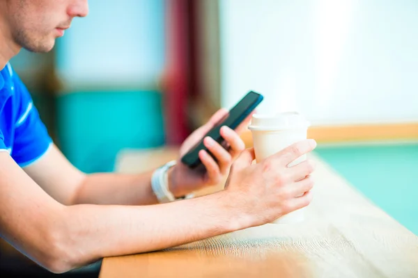Närbild av manliga händer som håller mobiltelefon och glas kaffe i caféet. Man använder mobiltelefon. Pojken vidrör en skärm av sin smarthone. suddig bakgrund, horisontell. — Stockfoto