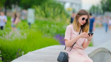 Beyaz bir kadın mesaj gönderiyor ve Avrupa 'da açık havada müzik dinliyor. Güneş gözlüklü güzel kız ahşap bankta akıllı telefon kullanarak oturuyor.