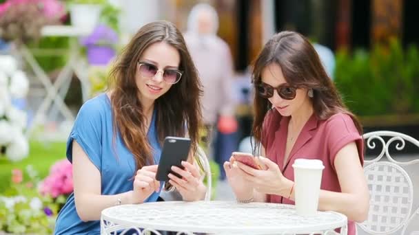 Dos chicas jóvenes usando un teléfono inteligente en la cafetería al aire libre. Dos mujeres después de ir de compras con bolsas sentadas en la cafetería al aire libre con café y el uso de smartphone — Vídeos de Stock