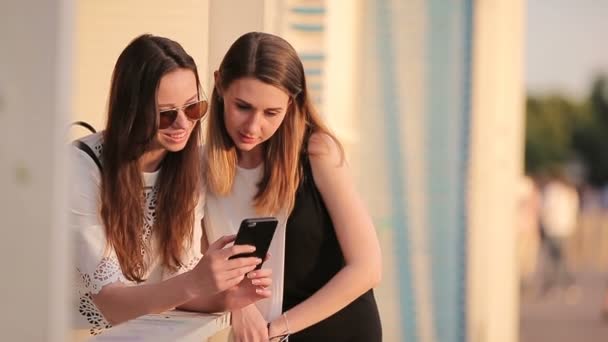 Δύο νεαρά κορίτσια χρησιμοποιώντας έξυπνο τηλέφωνο σε εξωτερικούς χώρους. Δύο γυναίκες συνεδρίαση στο πάρκο στο φως του ηλιοβασιλέματος και χρησιμοποιώντας smartphone — Αρχείο Βίντεο