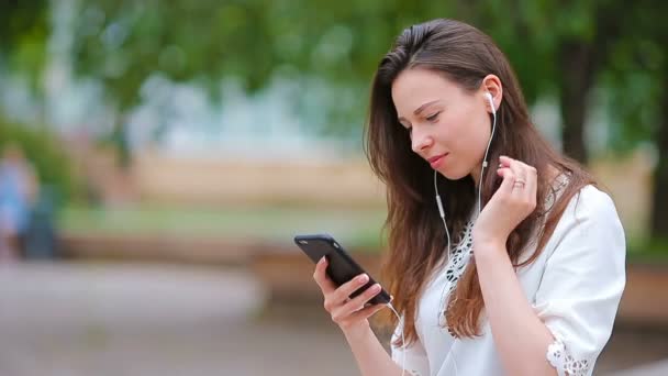 Jonge Kaukasische vrouw verzenden van bericht en luisteren muziek buiten op Europese stad. Mooi meisje zittend op houten bankje smartphone met zacht zonsondergang licht — Stockvideo