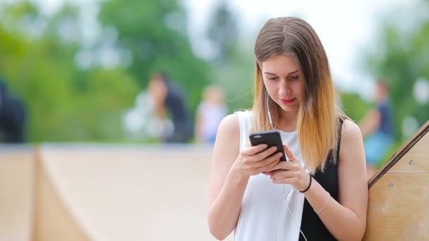 Junge kaukasische Frau sendet Botschaft und hört Musik im Freien in der europäischen Stadt. Schönes Mädchen mit Sonnenbrille sitzt mit Smartphone auf Holzbank — Stockvideo