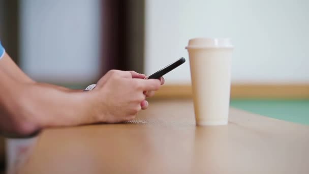 Close-up van mannelijke handen met mobiele telefoon en een glas koffie in het café. Man met mobiele smartphone. Jongen die een scherm van zijn slimmerik aanraakt. Wazige achtergrond, horizontaal. — Stockvideo