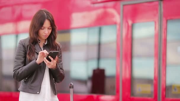 Mujer joven con equipaje mirando oh teléfono celular en una estación de tren. Caucasiam turista esperando su expreso tren mientras su viaje . — Vídeo de stock