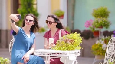 Açık hava kafesinde iki genç kız akıllı telefonlarıyla selfie çekiyor. Çantalarla alışveriş yaptıktan sonra açık kafede kahve içip akıllı telefon kullanan iki kadın.