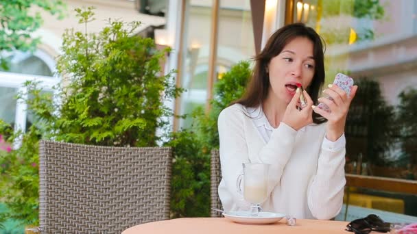 Mooie vrouw haar lippen verven met rode lippenstift op terras zitten. Jong meisje met heerlijke zoete cappuccino. — Stockvideo