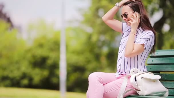 Giovane ragazza caucasica parlando dal suo smartphone all'aperto. Bella ragazza in occhiali da sole per strada utilizzando smartphone e parlando per telefono con la sua amica — Video Stock