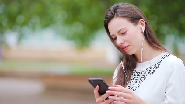 Νεαρή καυκάσια γυναίκα στέλνει μήνυμα από το smartphone της στο πάρκο. Όμορφο κορίτσι με γυαλιά ηλίου κάθεται σε ξύλινο πάγκο χρησιμοποιώντας smartphone — Αρχείο Βίντεο
