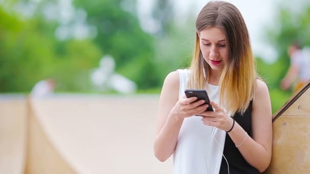 Mujer caucásica joven que envía mensajes y escucha música al aire libre en la ciudad europea. Hermosa chica en gafas de sol sentado en el banco de madera con el teléfono inteligente — Vídeo de stock