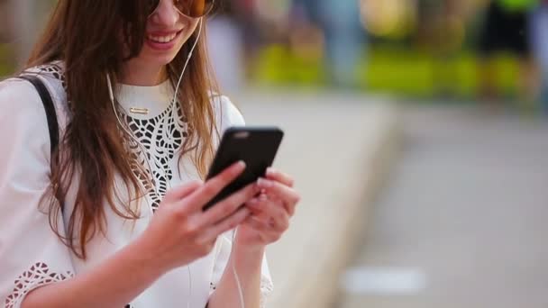 Mujer caucásica joven que envía un mensaje por su teléfono inteligente al aire libre en el parque al atardecer. Hermosa chica en gafas de sol sentado en el banco de madera con el teléfono inteligente — Vídeo de stock