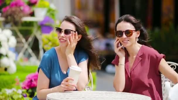 Dos chicas jóvenes usando un teléfono inteligente en la cafetería al aire libre. Dos mujeres después de ir de compras con bolsas sentadas en la cafetería al aire libre con café y el uso de smartphone — Vídeos de Stock