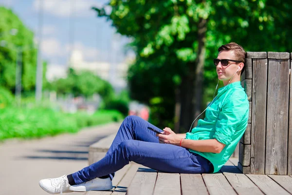 Jongen luisteren muziek via smartphone op zomer vakantie. Jonge aantrekkelijke toerist met mobiele telefoon buitenshuis genieten van vakantie. — Stockfoto