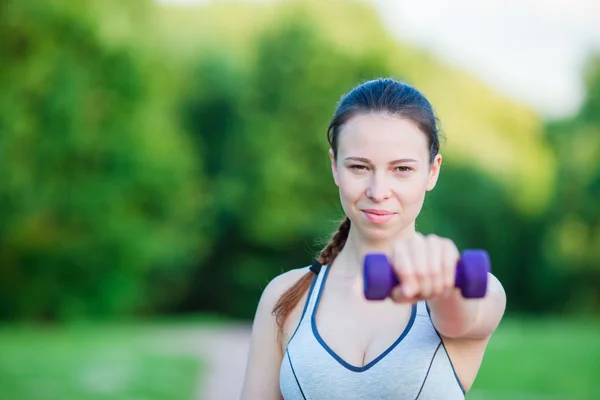 Ajuste mujer joven que trabaja con pesas al aire libre. Chica activa haciendo ejercicio con pequeñas mancuernas en el parque — Foto de Stock