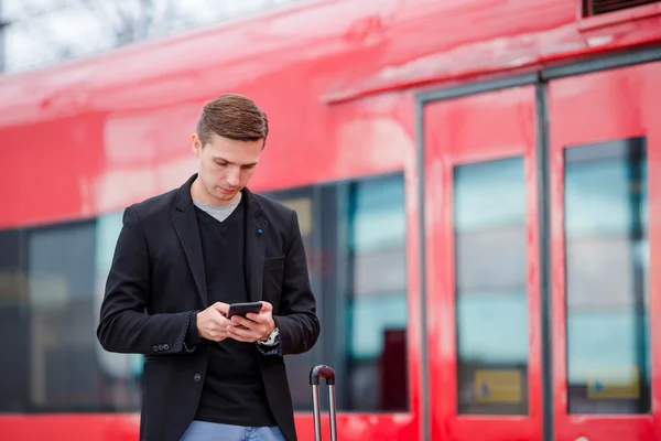 Jovem caucasiano com smartphone e bagagem na estação viajando de trem — Fotografia de Stock