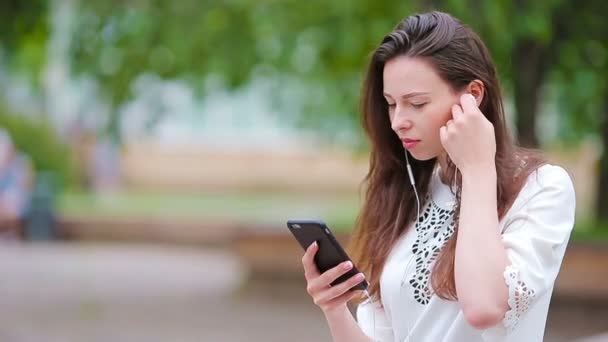 Jonge Kaukasische vrouw stuurt bericht via haar smartphone buiten in het park. mooi meisje in zonnebril zitten op houten bank met behulp van smartphone — Stockvideo