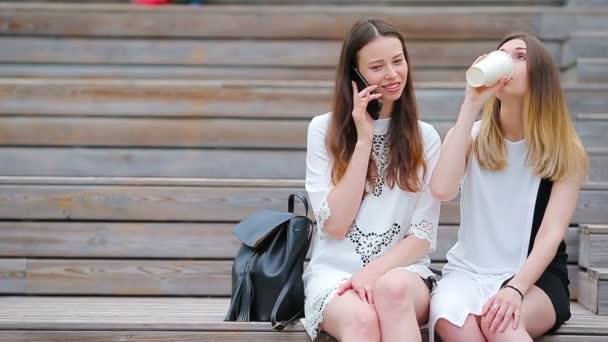 Dwie młode dziewczyny używające smartfona na zewnątrz. Dwie kobiety siedzące w parku z kawą i korzystające ze smartfona — Wideo stockowe