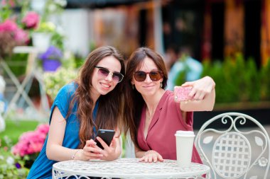 Açık hava kafesinde iki genç kız akıllı telefonlarıyla selfie çekiyor. Çantalarla alışveriş yaptıktan sonra açık kafede kahve içip akıllı telefon kullanan iki kadın.