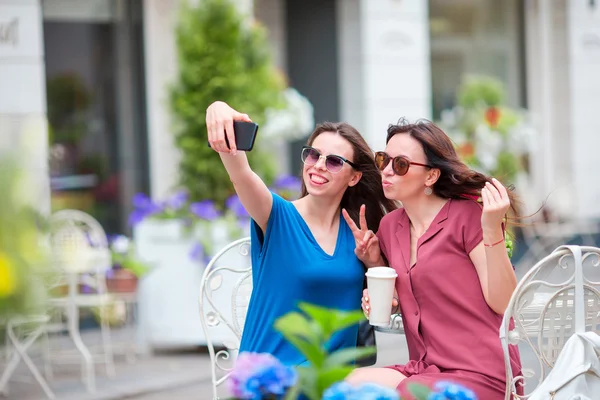 Two young girls taking selfie with smart phone at the outdoors cafe. Two women after shopping with bags sitting in openair cafe with coffee and using smartphone — Stock Photo, Image